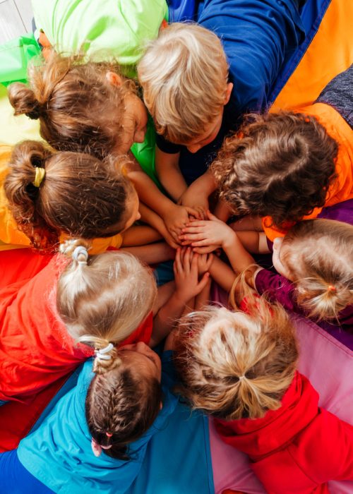 Lovely children laying on multicolor canopy in circle. Team building game for daycare children in colorful t-shirts. Top view kids on wooden floor.